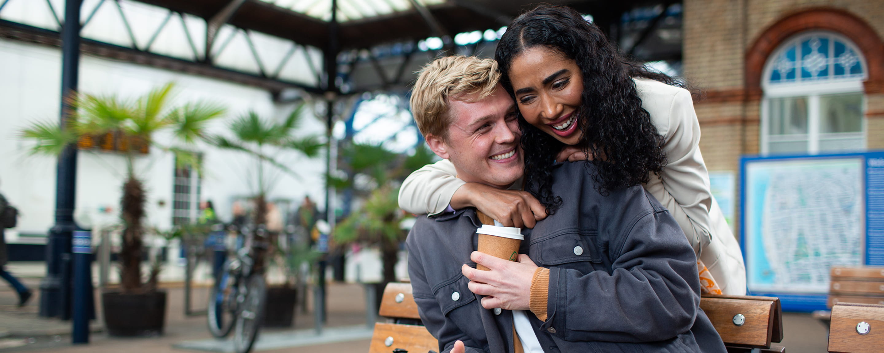 A woman smiling at a man sitting on a bench at a railway station