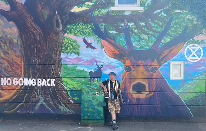 A man standing on a street, leaning against a wall with a large mural painting