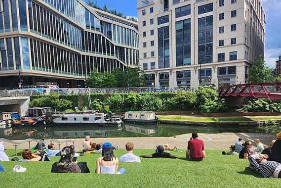 People sitting on the grassy bank of a river with long boats and buildings in the background